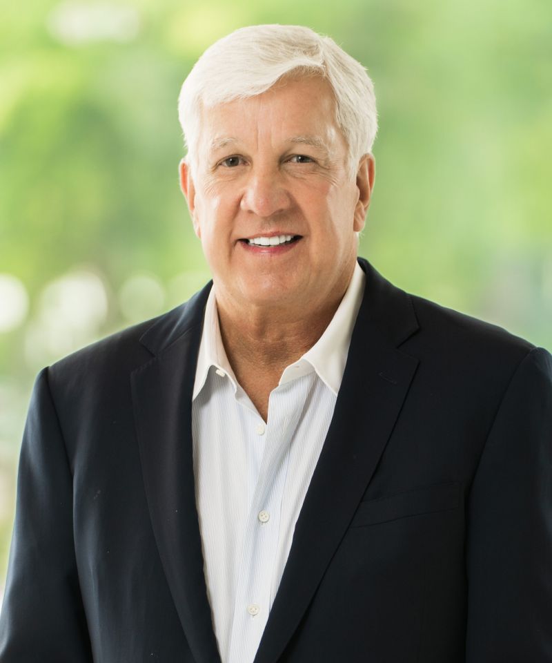 Smiling older man in a dark suit jacket and white shirt, standing in front of a blurred green background.
