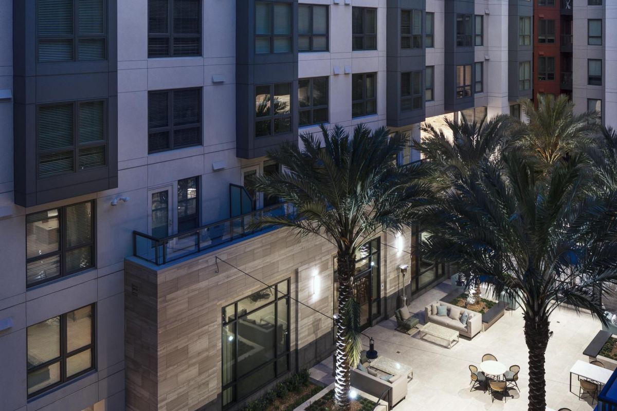 Modern apartment courtyard at dusk with palm trees, fountain, tables, chairs, and lit pathways.