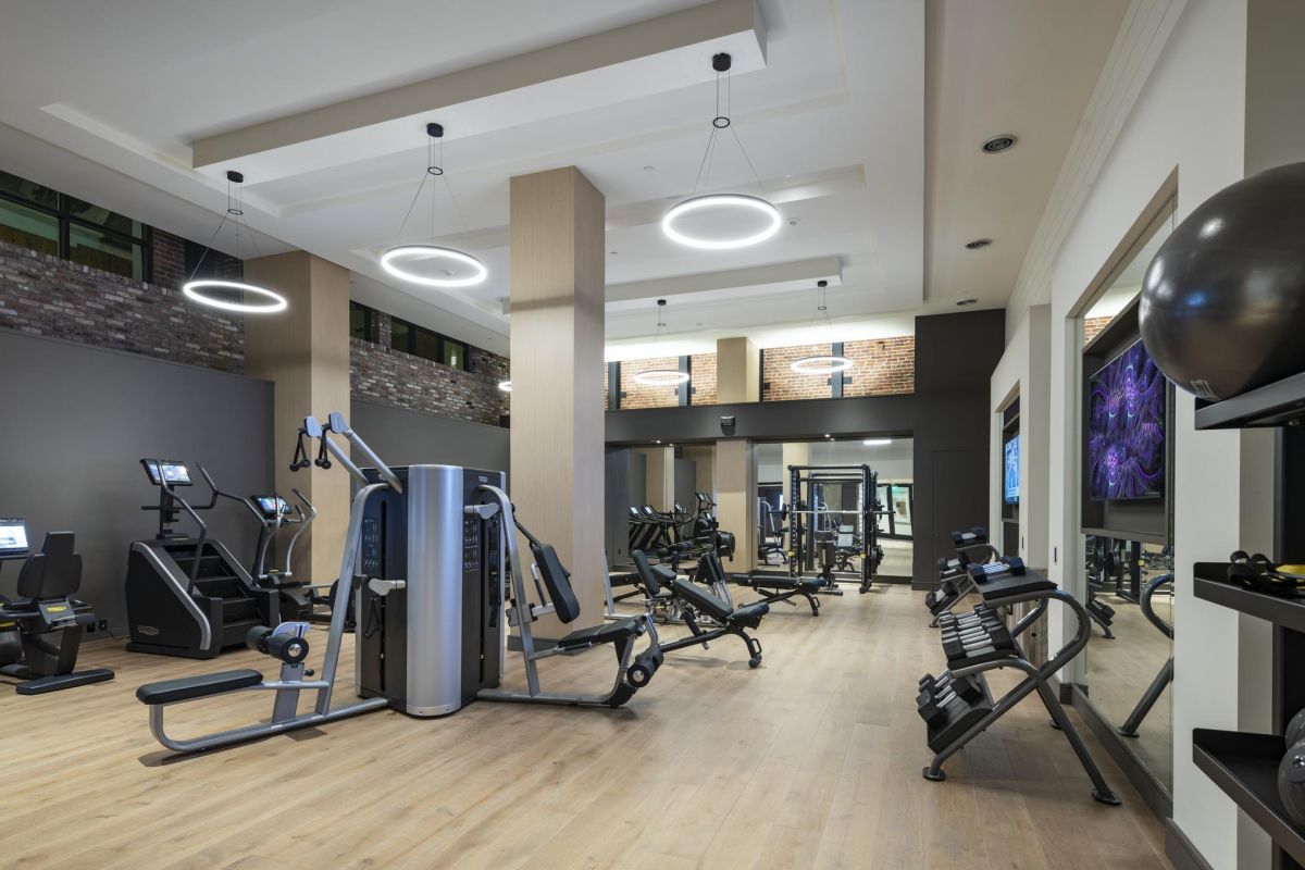 Modern gym with exercise machines, large mirrors, wooden flooring, and circular ceiling lights.
