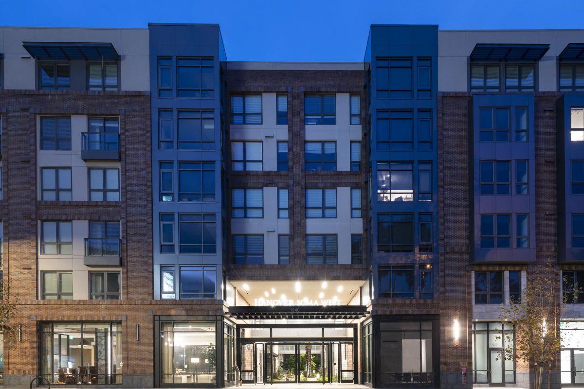 Illuminated modern apartment building at dusk with glass entrance and trees lining the sidewalk.