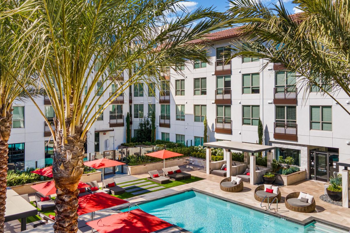 Modern apartment complex with palm trees, a pool, red umbrellas, and lounge areas in a courtyard setting.