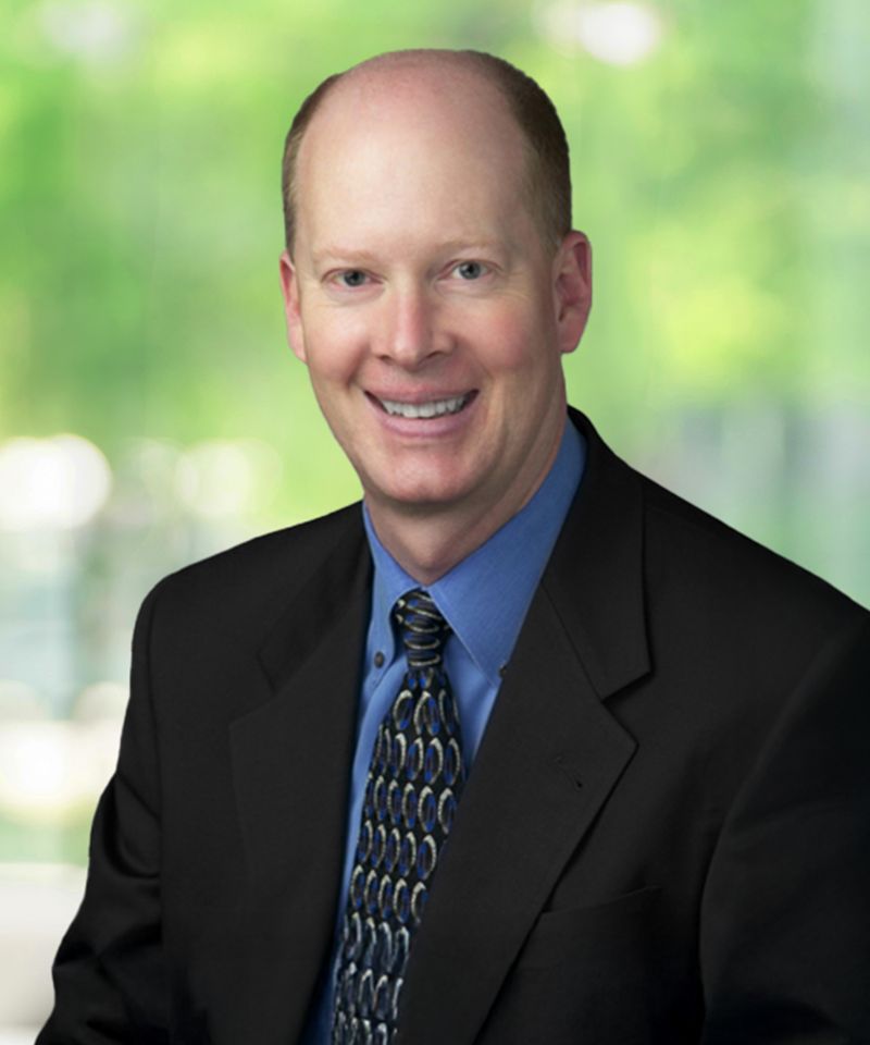 Smiling man in a suit and blue shirt, seated with a blurred green background.
