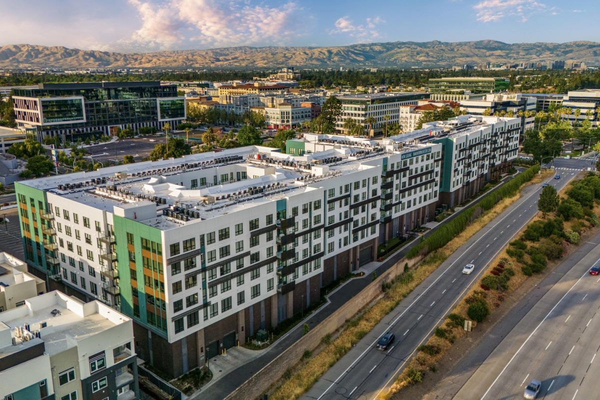 Aerial view of urban buildings with a highway nearby, mountains, and a cloudy sky in the background.