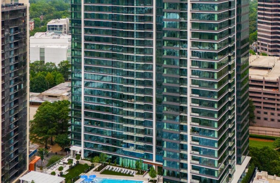 Tall glass apartment building with a pool and trees at its base, surrounded by other high-rise buildings.
