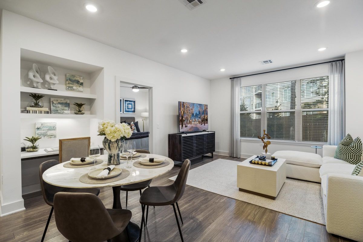 Modern living and dining area with a round table, chairs, white sofa, TV, large window, and decorative shelves.