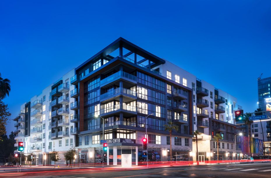 Modern apartment building at dusk with illuminated windows, streetlights, and light trails from passing cars.