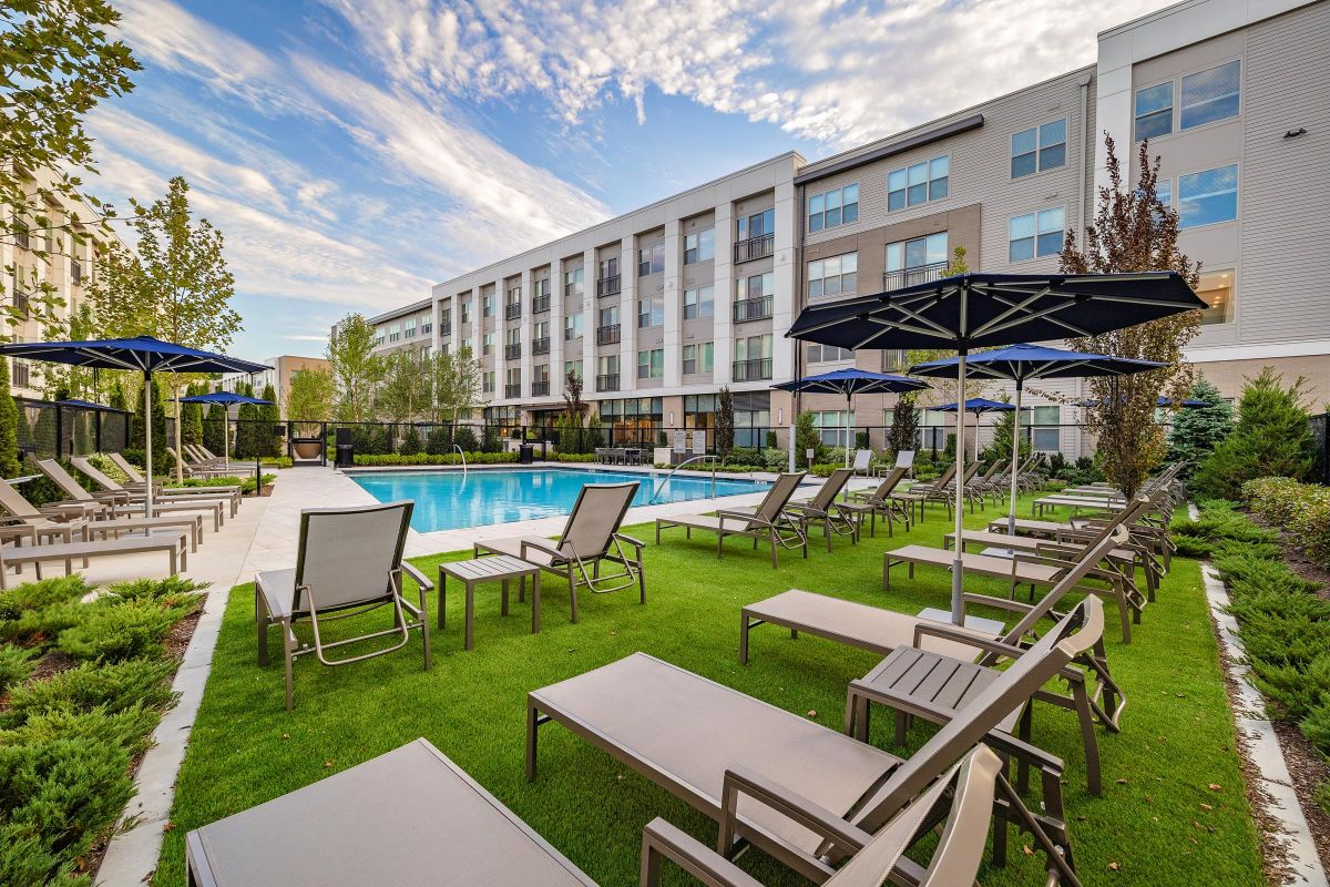 Outdoor pool area with lounge chairs and umbrellas, surrounded by modern apartment buildings and greenery under a cloudy sky.