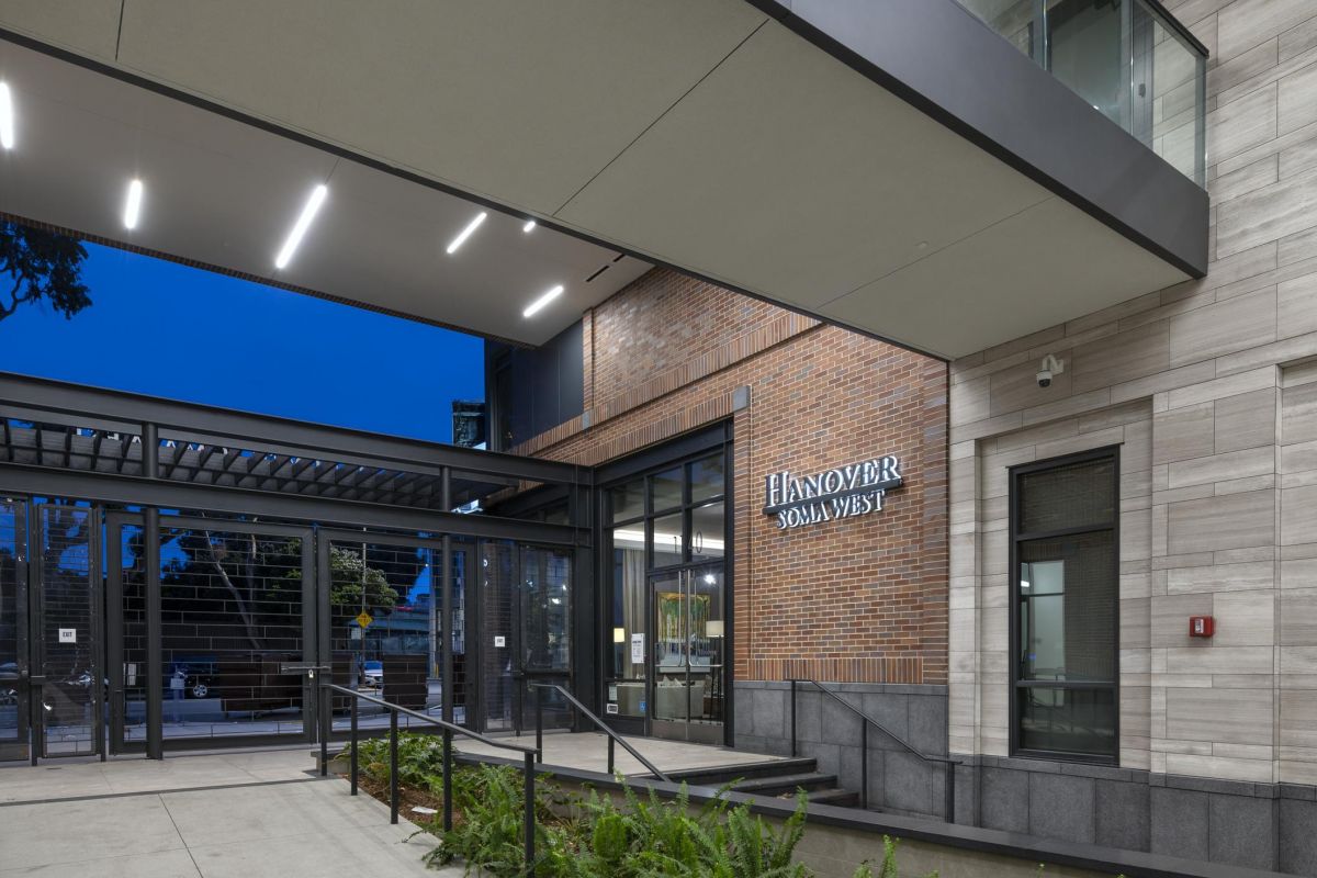Modern building entrance with glass doors, tall plants, and "Hanover Solamar" sign on brick wall at dusk.