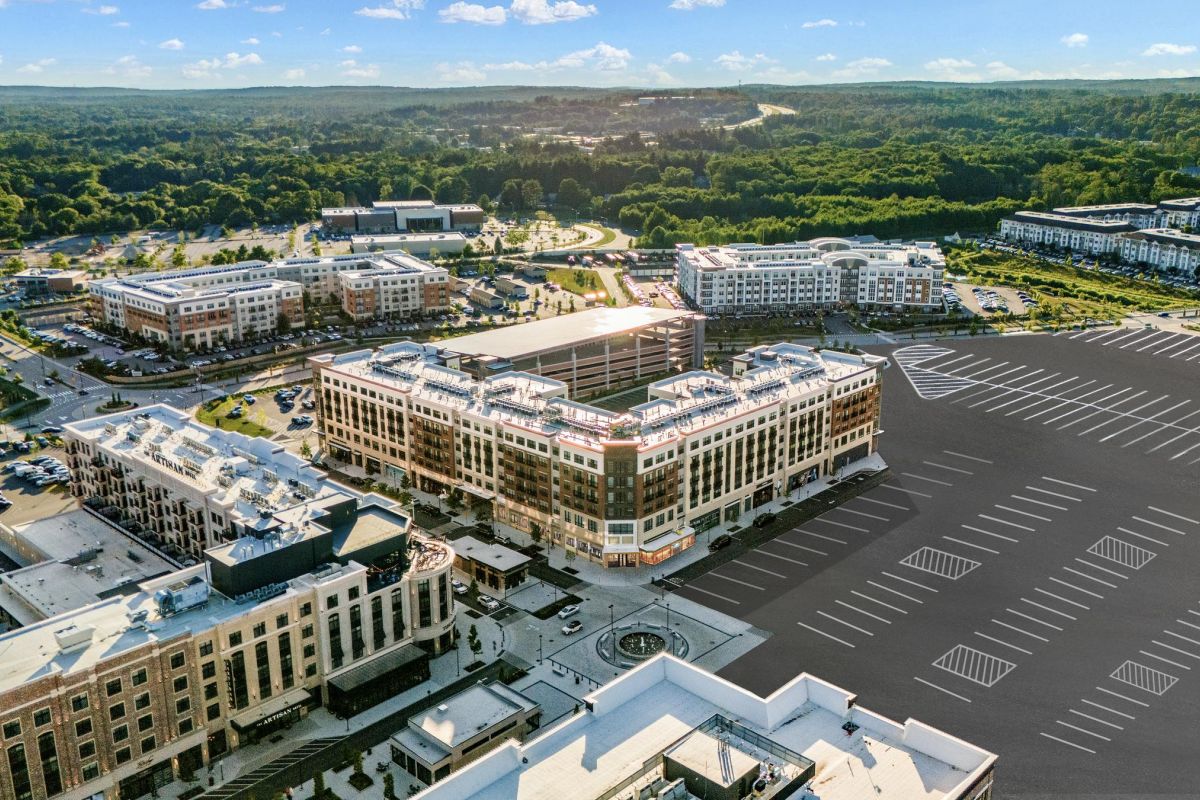 Aerial view of a large shopping center with multiple buildings and a nearly empty parking lot surrounded by greenery.