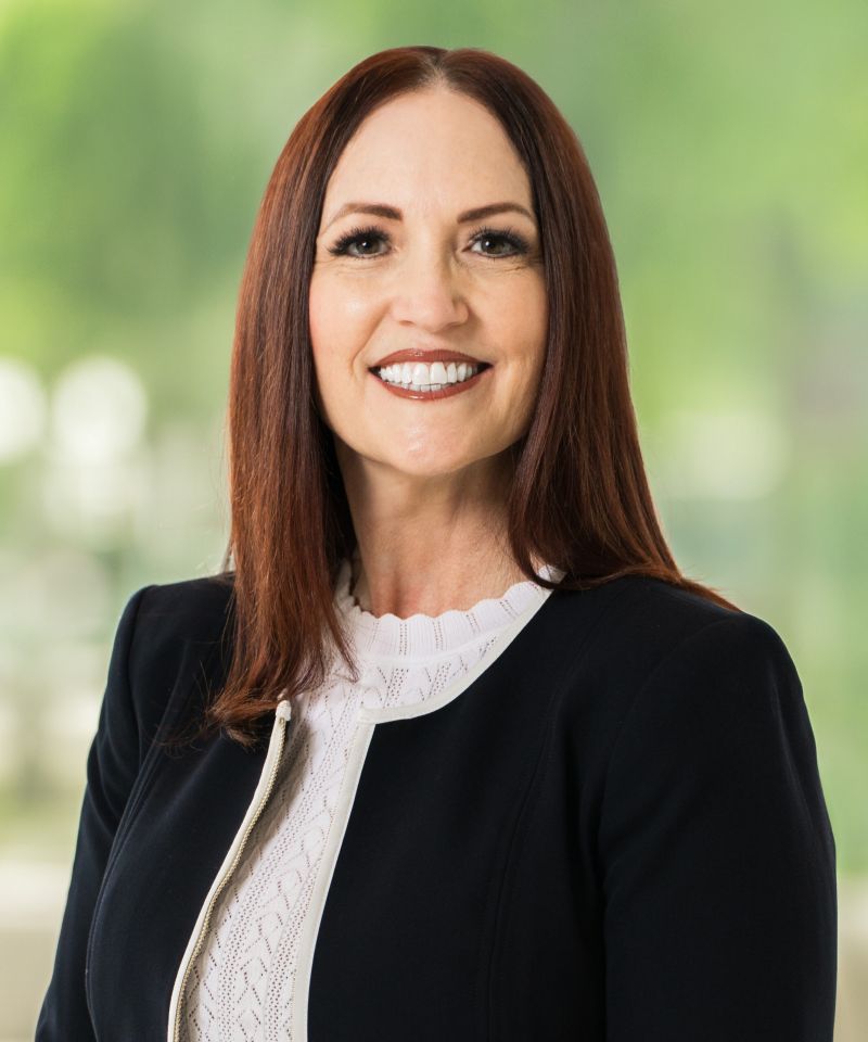 Smiling woman with long brown hair, wearing a black blazer and white top, standing in front of a blurred background.