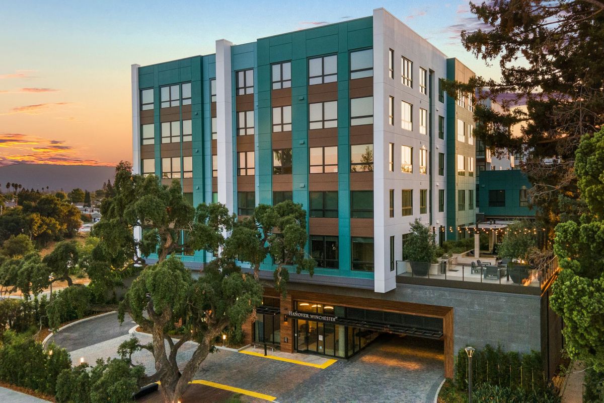 Modern five-story hotel building with large windows, surrounded by trees, at sunset.