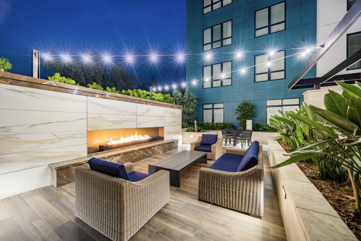 Outdoor seating area with wicker furniture, a modern fireplace, string lights above, and plants around at night.