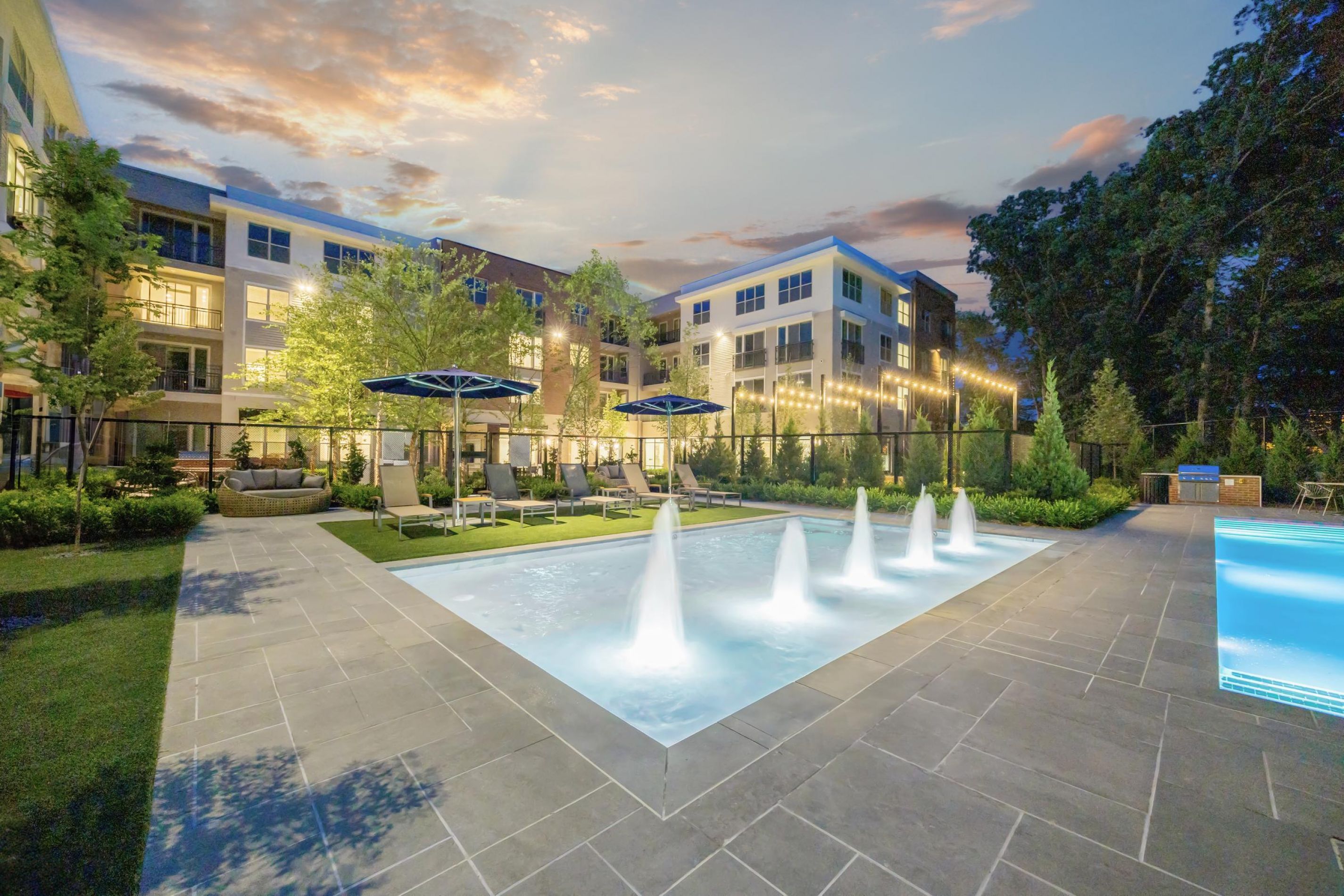 Modern apartment complex with a pool, fountains, and string lights at dusk, surrounded by trees and greenery.