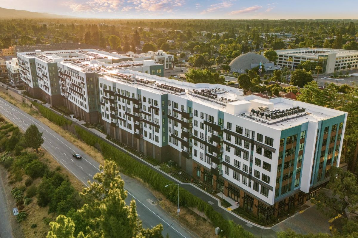 Aerial view of a modern multi-story apartment building beside a road, surrounded by trees and cityscape under a blue sky.