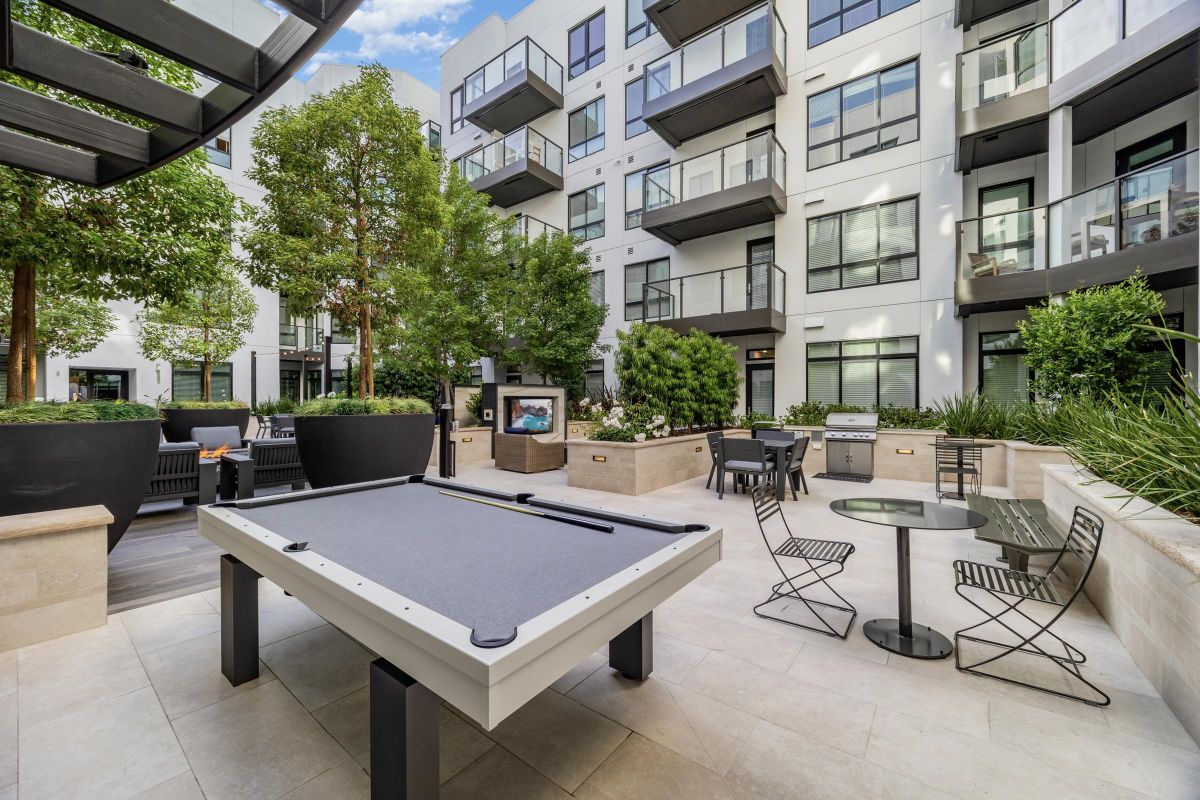 Outdoor patio with a pool table, seating, grill, and greenery in a modern apartment complex courtyard.