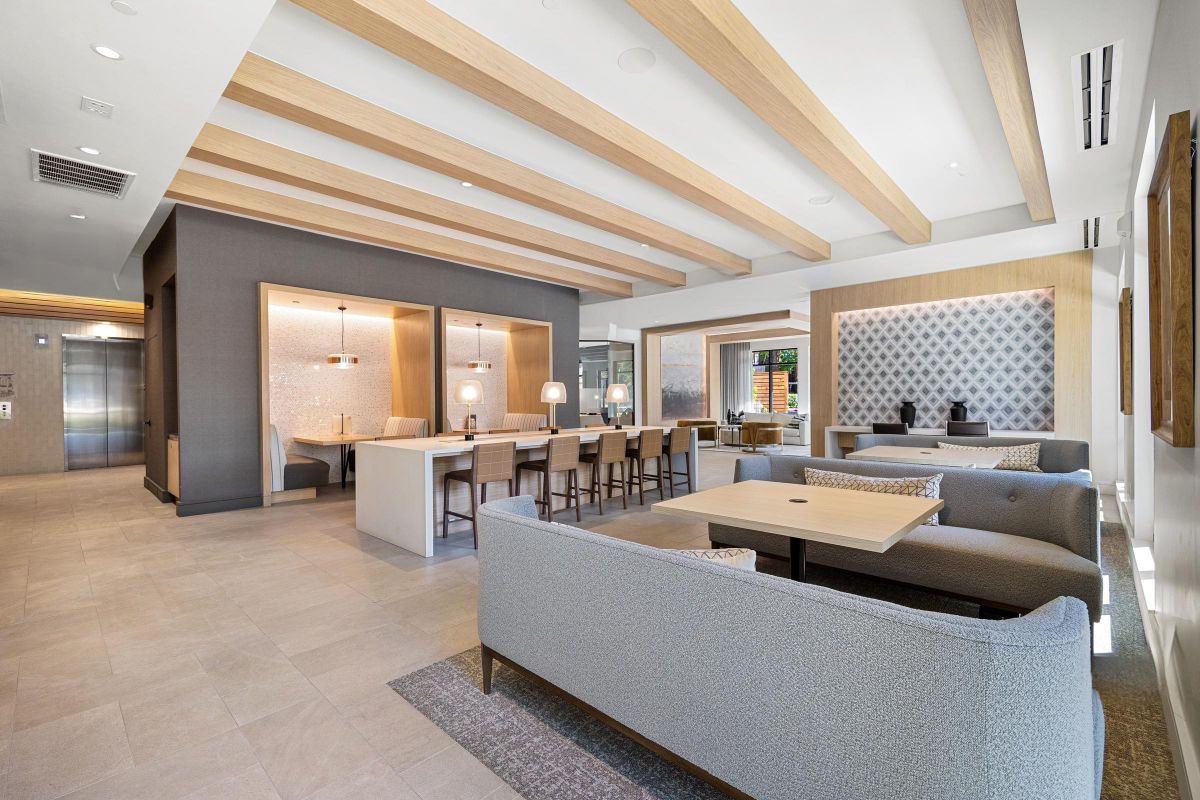 Modern lounge with gray sofas, wooden beams, a long bar table, and neutral-toned decor. Bright and spacious setting.