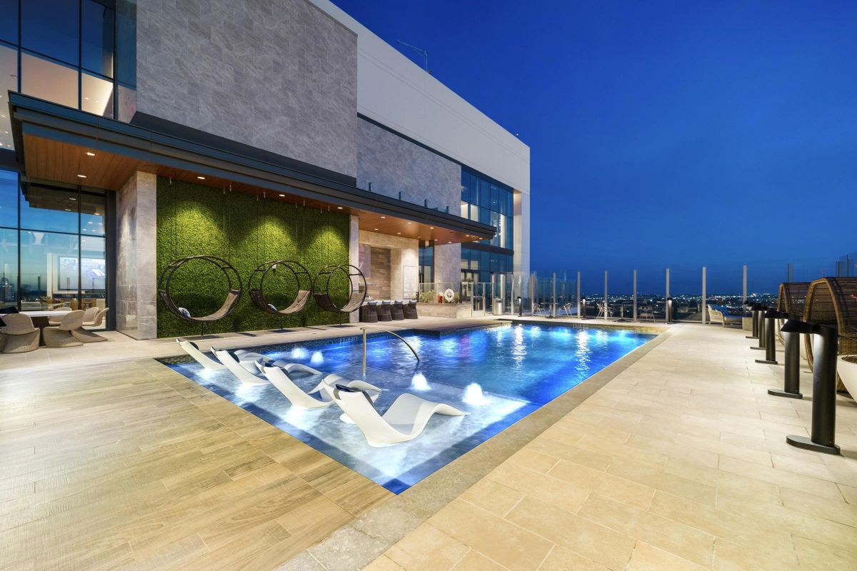 Rooftop pool at night with lounge chairs and circular swings, overlooking city lights and a glass barrier.