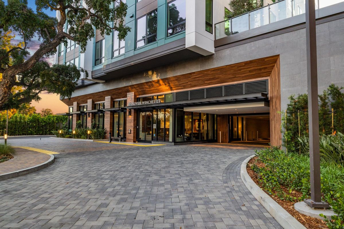 Modern hotel entrance with large windows, wooden accents, and a paved driveway surrounded by landscaped greenery.