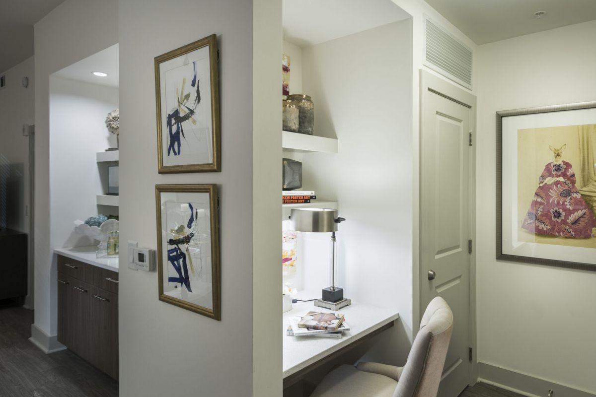 Small home office nook with a desk, chair, artwork, and shelves, adjacent to a hallway and kitchen.