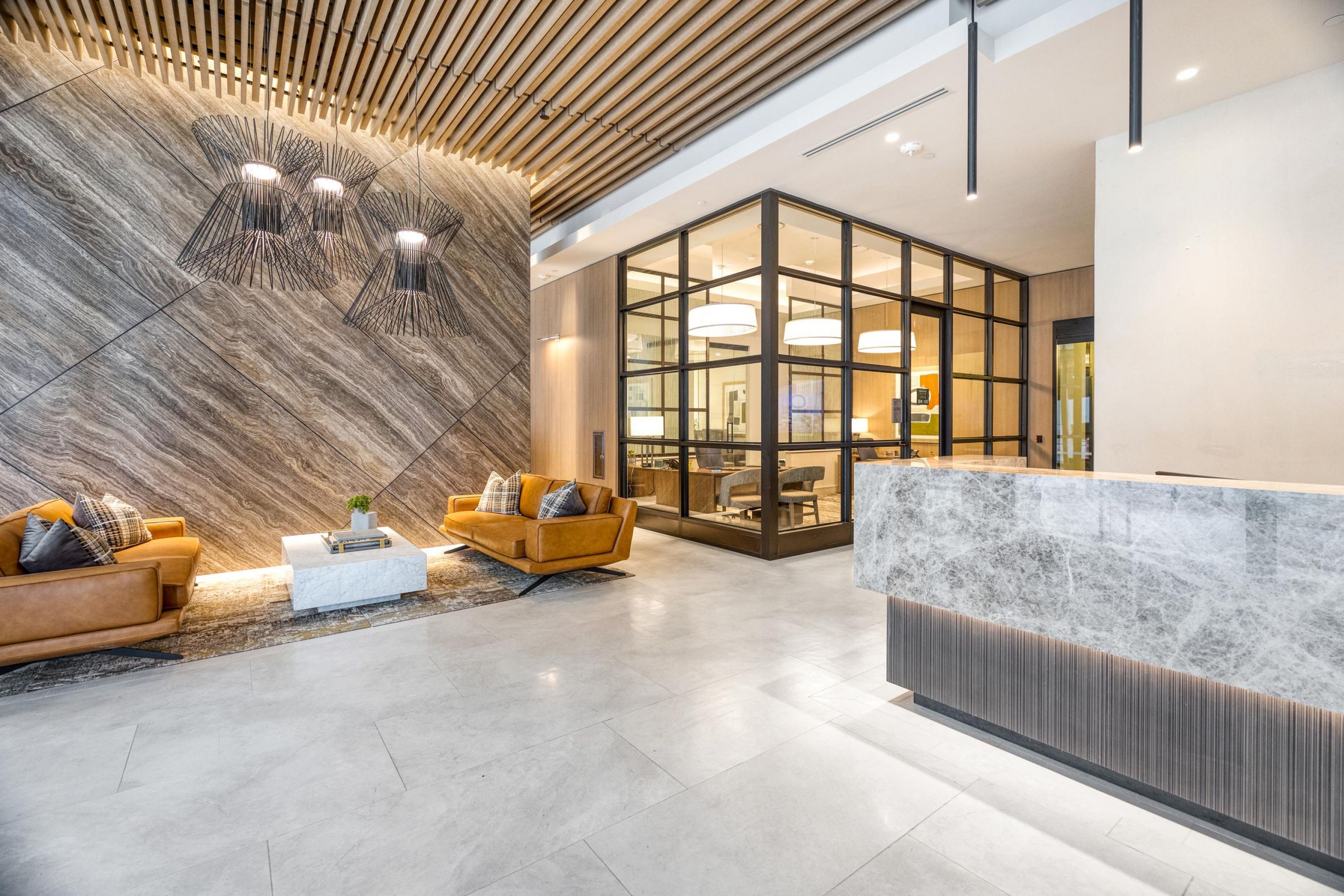 Modern lobby with marble reception desk, tan sofas, and glass-walled meeting room, featuring elegant lighting.