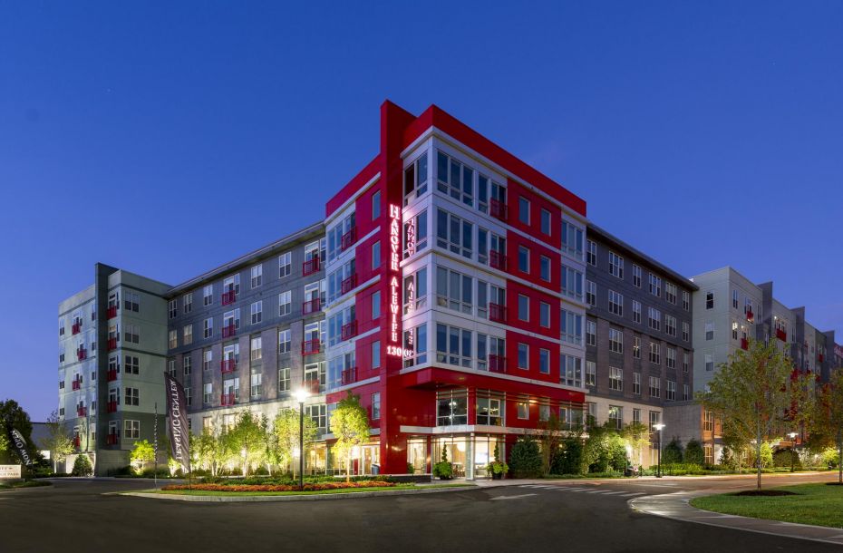 Modern apartment building with red accents, surrounded by greenery, lit up at dusk against a clear blue sky.