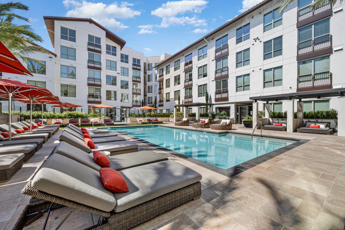 Outdoor pool area with lounge chairs, red umbrellas, and modern apartment buildings in the background.