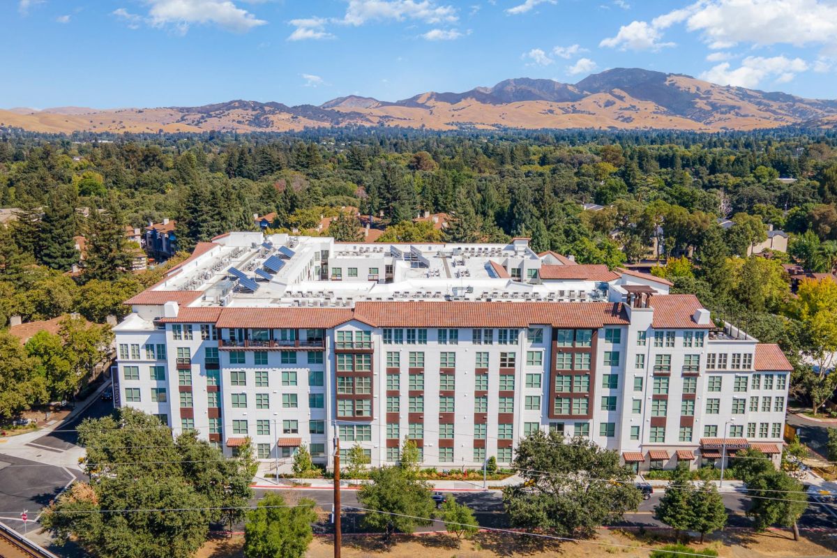 Large white building surrounded by trees, with mountains in the background under a blue sky.