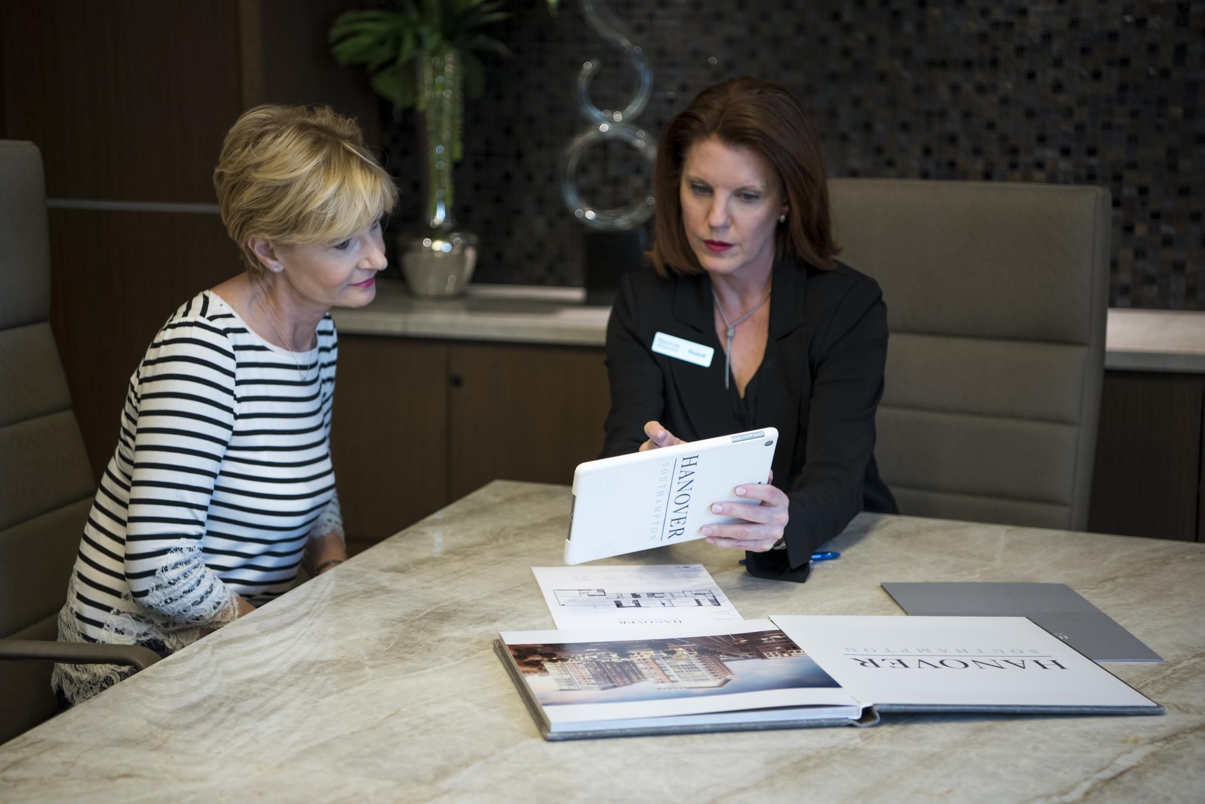 Two women sit at a table, one showing the other a tablet with brochures and papers laid out.