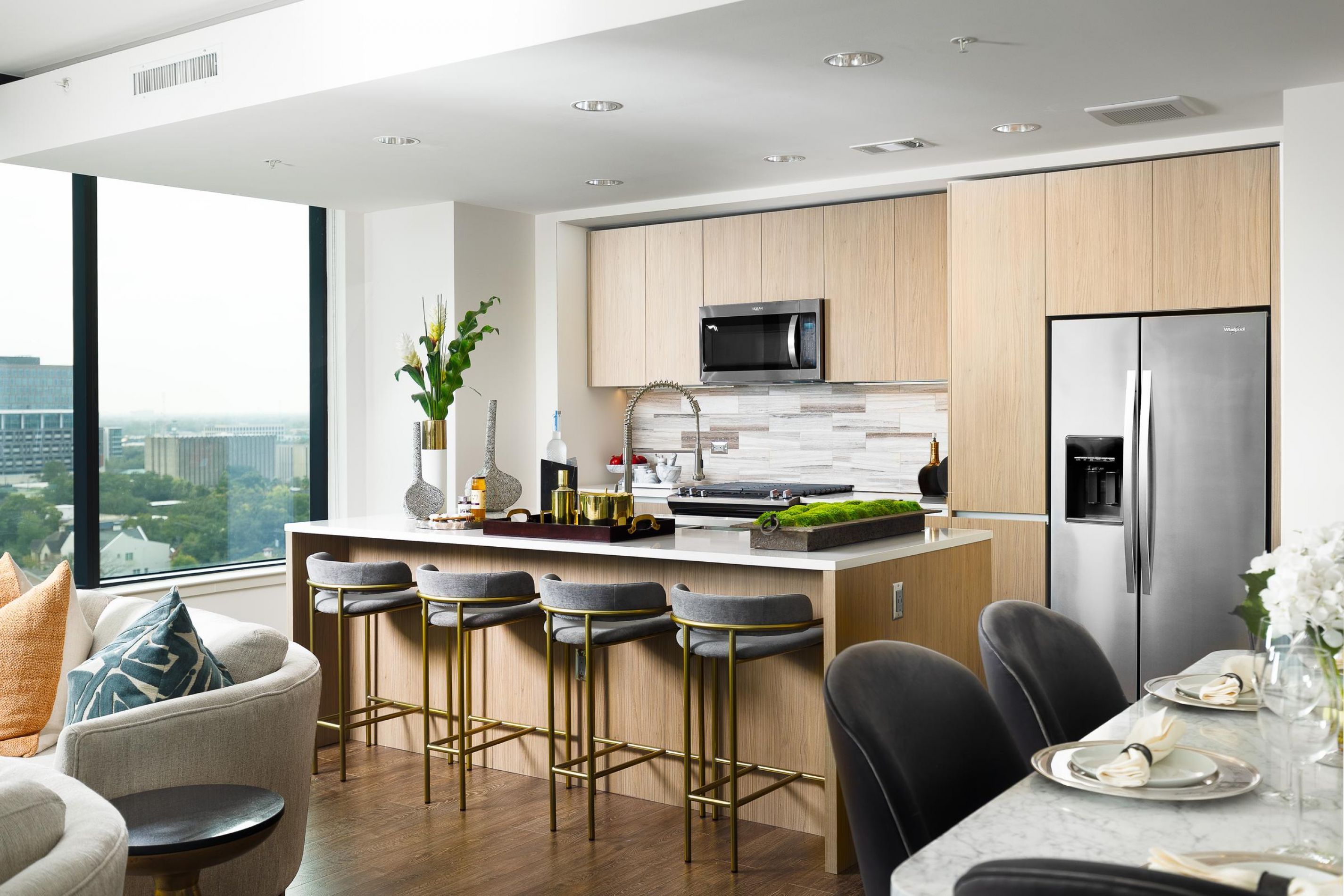 Modern kitchen with wood cabinets, bar stools, stainless steel appliances, and large windows overlooking a city view at Hanover River Oaks – Houston, TX