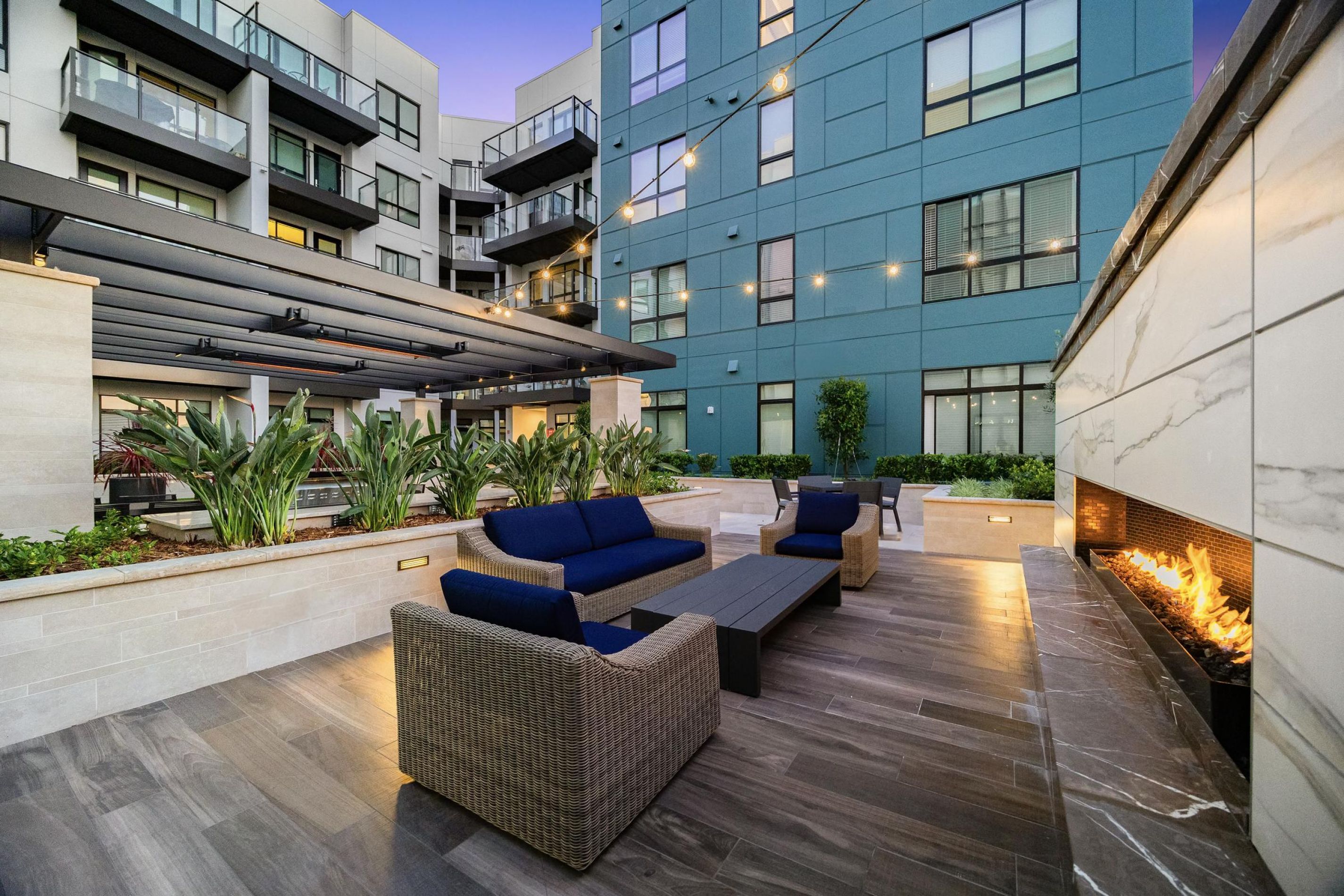 Modern outdoor patio with wicker sofas, a fire pit, and a pergola, surrounded by a tall building at dusk.