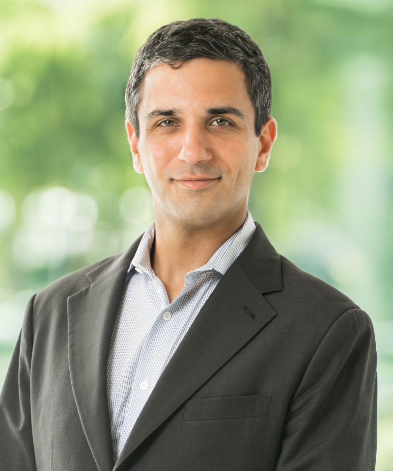 A man in a dark suit and striped shirt smiles, standing in front of a blurred green background.