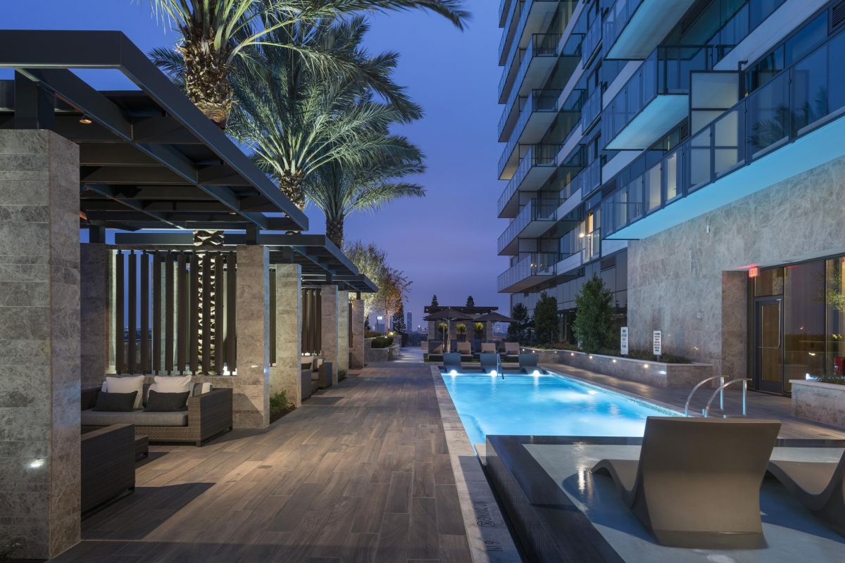 Modern poolside area at dusk, with lounge chairs, palm trees, and a lit apartment building in the background.