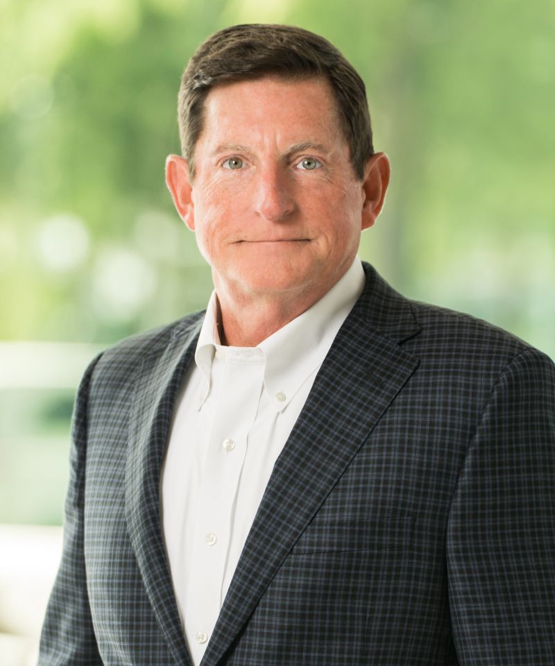 Rick Stinson, in a checkered suit jacket and white shirt, stands against a blurred green outdoor background.