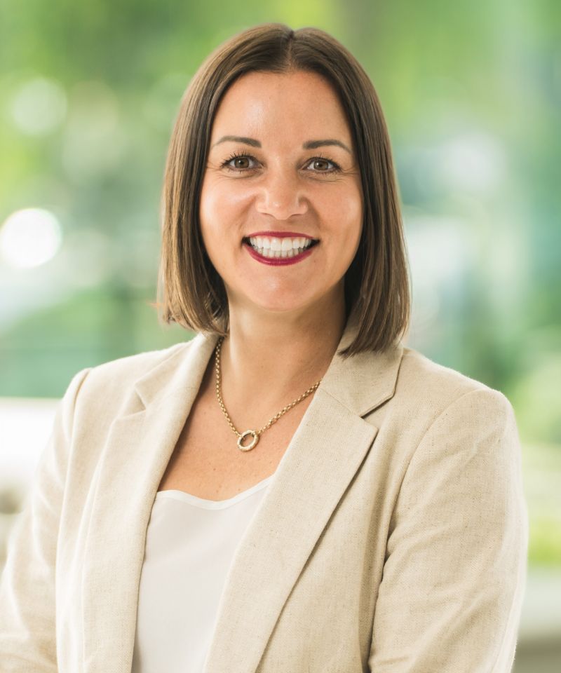 Deanna Chalfant with a bob haircut smiles, wearing a beige blazer and gold necklace, set against a blurred green background.
