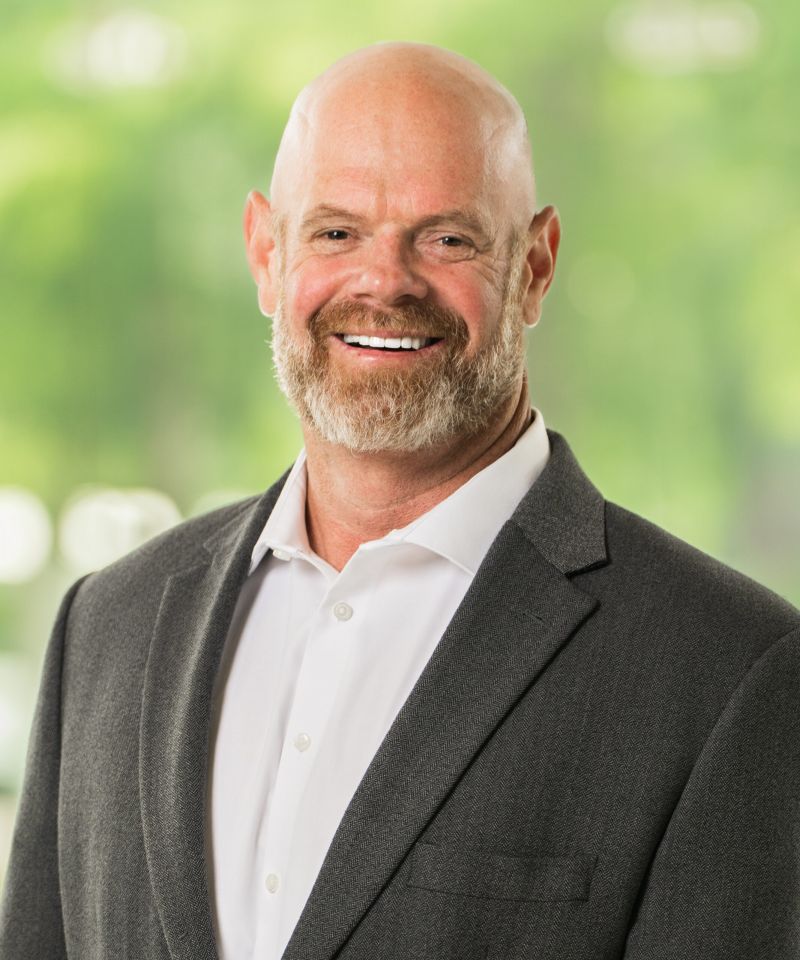 Smiling bald man with a beard in a gray suit jacket and white shirt, standing outdoors with greenery behind.