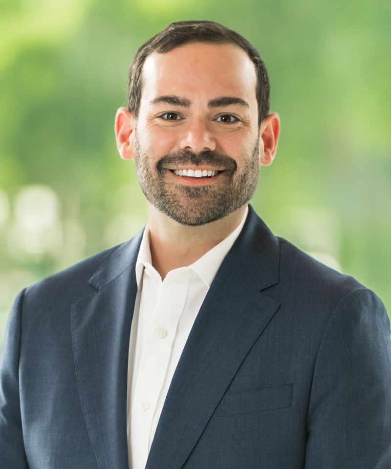 Smiling man in a navy suit and white shirt, standing in front of a blurred green background.