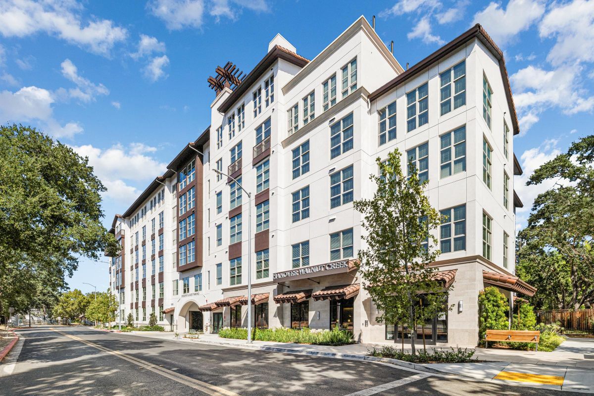 Modern multi-story building with large windows and street view, surrounded by trees under a blue sky.