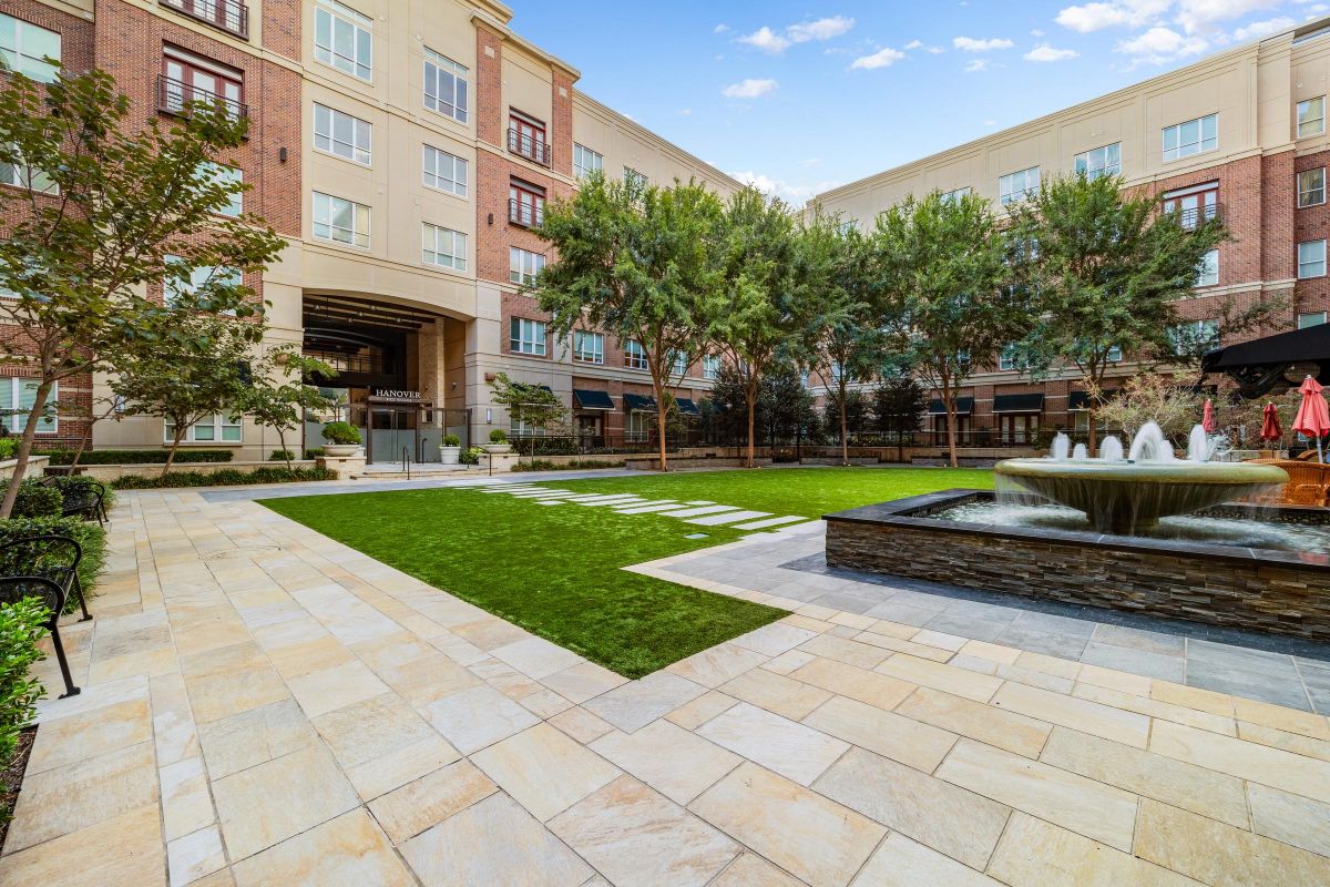 Hanover Company Courtyard with grass, a water fountain, trees, and a brick building in the background under a blue sky.