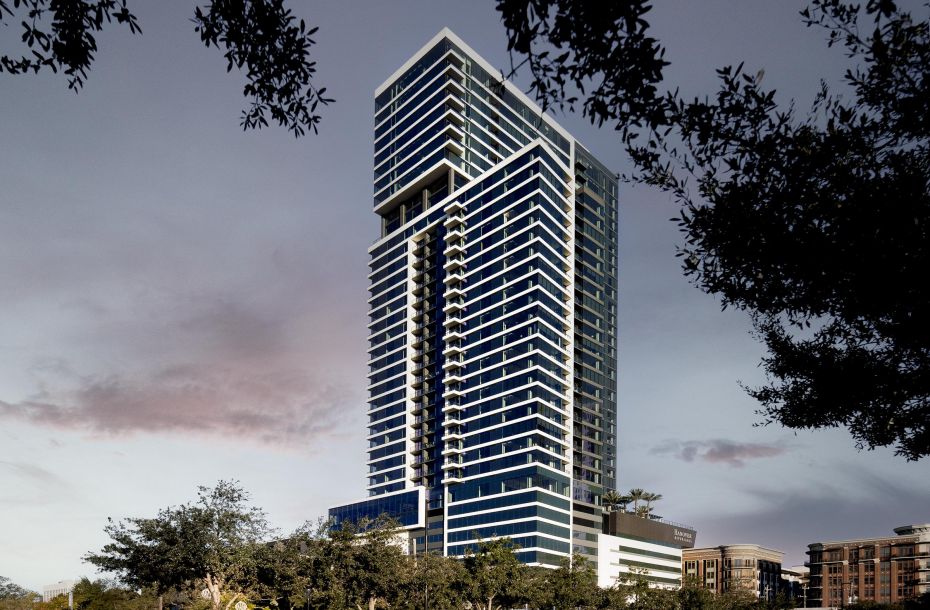 Modern high-rise building with glass facade, surrounded by trees, under a cloudy sky at dusk.