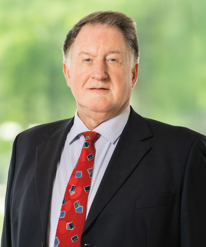 A man in a suit and red patterned tie stands in front of a blurred green background.