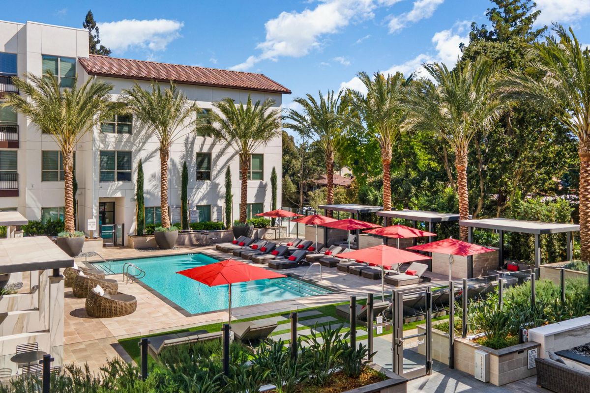 Modern apartment complex with a pool, red umbrellas, palm trees, and lounge chairs under a partly cloudy sky.