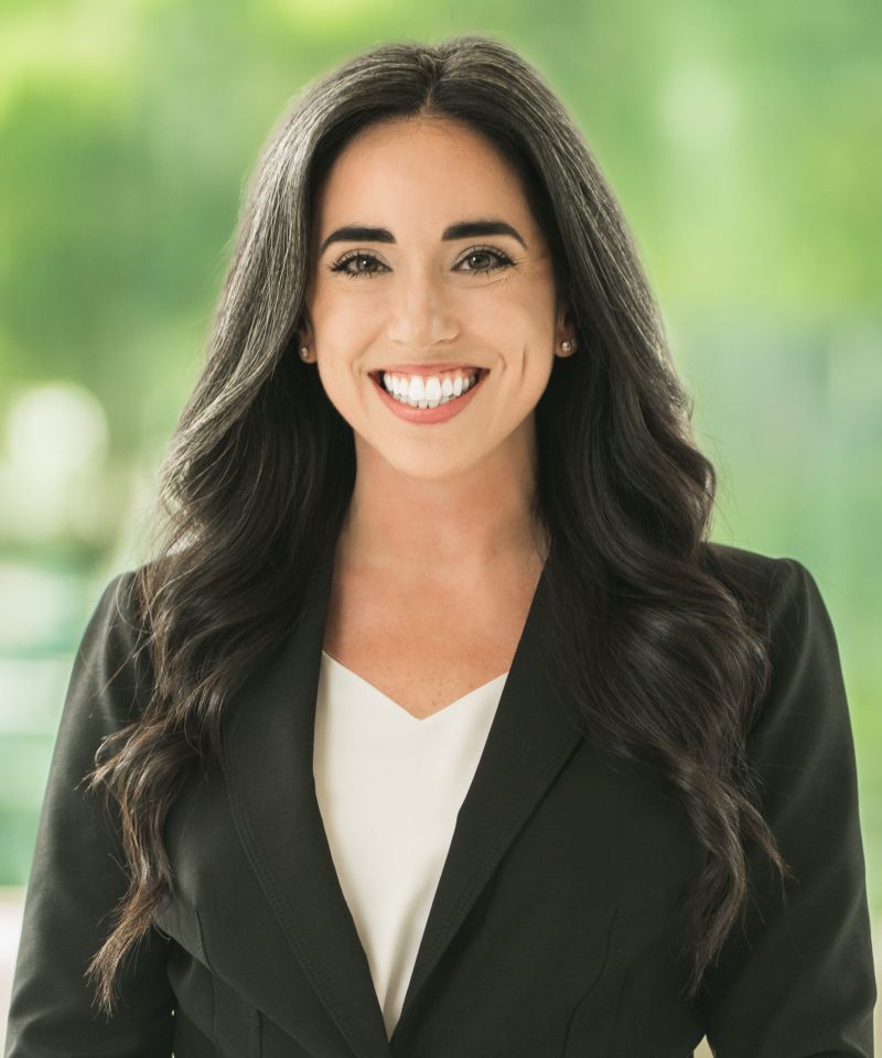Smiling woman with long dark hair in a black blazer, standing in front of a green blurred background.