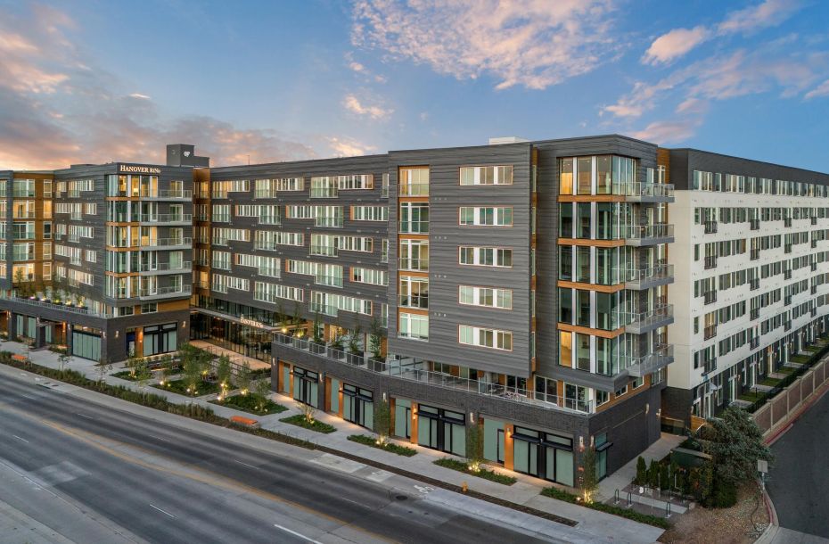 Modern multi-story apartment building with balconies, large windows, and street view under a blue sky with clouds.