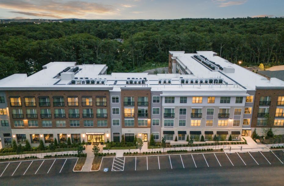 Aerial view of a modern apartment building with surrounding parking and forest in the background at sunset.