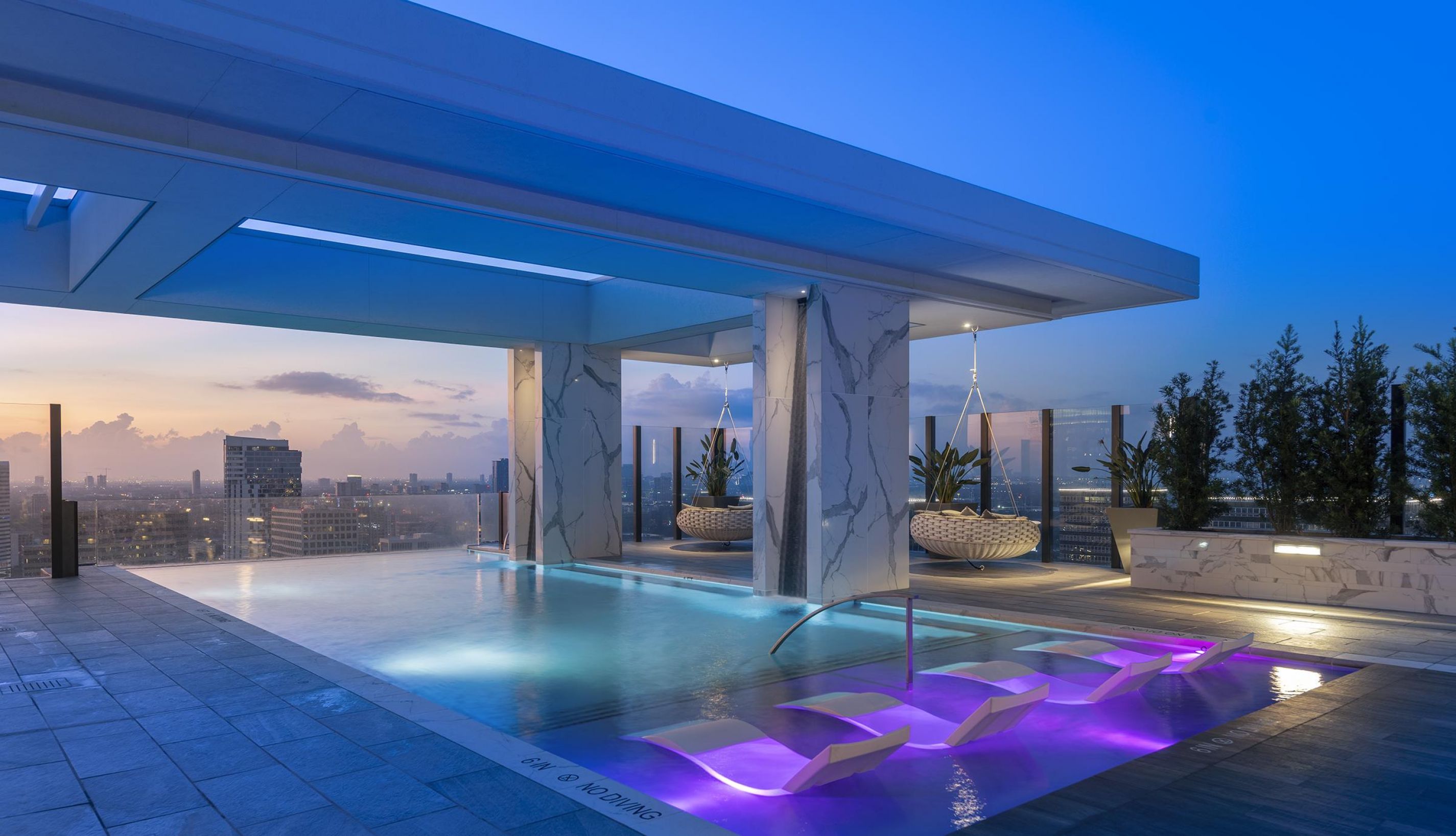 Rooftop infinity pool at dusk with city skyline view, lounge chairs, vibrant lighting, and modern architectural design at Hanover BLVD Place – Houston, TX.