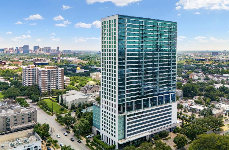 Aerial view of a modern high-rise building surrounded by trees and urban landscape under a partly cloudy sky.