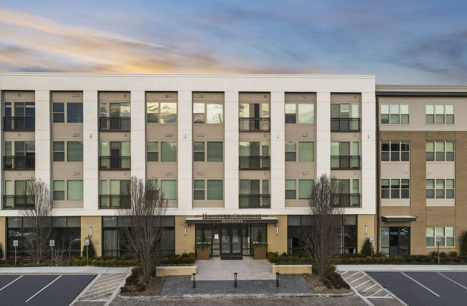 Hanover Company, Hanover Crossing Residences Modern four-story apartment building with large windows, at sunset. Empty parking lot in the foreground.