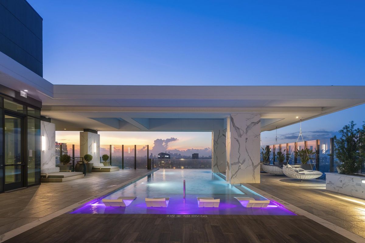 Modern rooftop pool area at sunset with lounge chairs, glass railings, and city skyline in the background.
