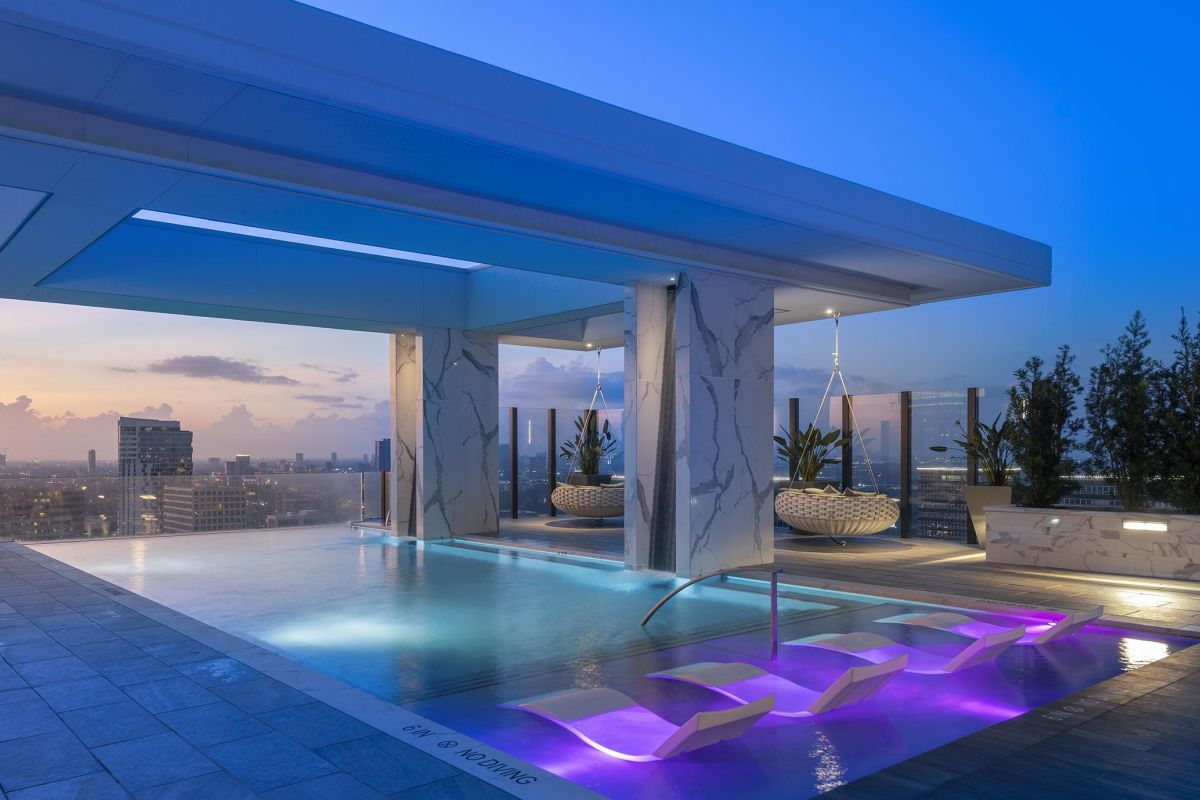 Rooftop infinity pool at sunset, with city skyline, lounge chairs in water, and modern cabanas on the deck.