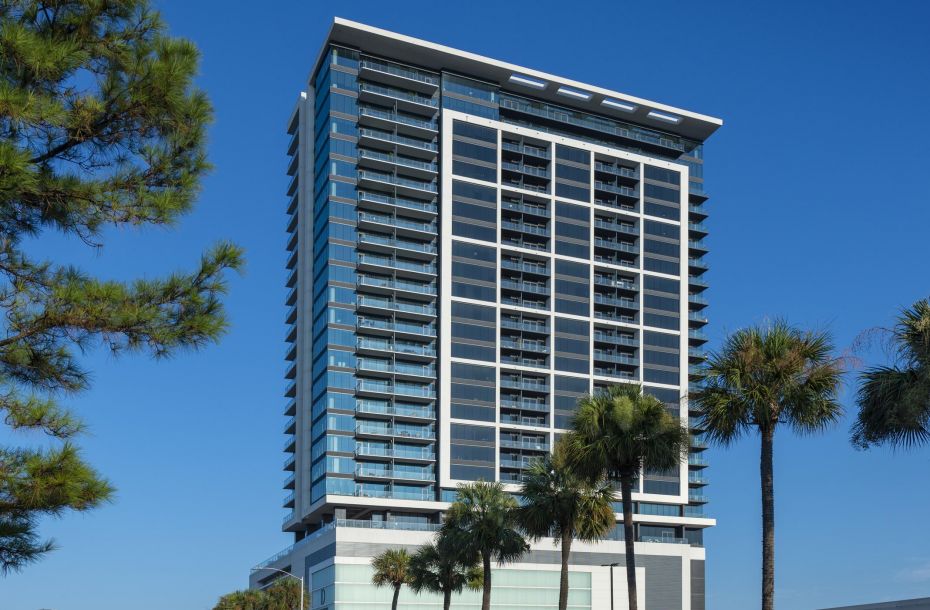 Tall modern building with glass facade, surrounded by palm trees and a clear blue sky. Street view with parked cars.